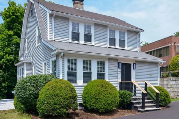 Two-story white colonial house with dark roof, black shutters, brick chimney, and manicured boxwood shrubs in front yard.